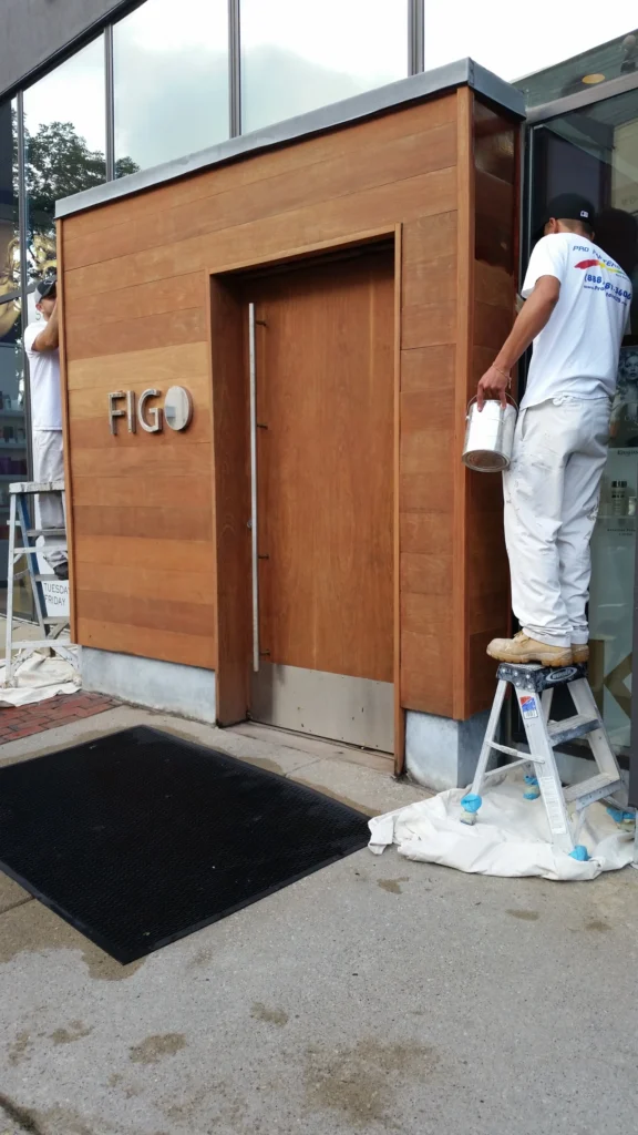 Pro Painters team preparing and refinishing the exterior wood entrance at FIGO, with drop cloths, ladders and protective masking in place during the finishing process.