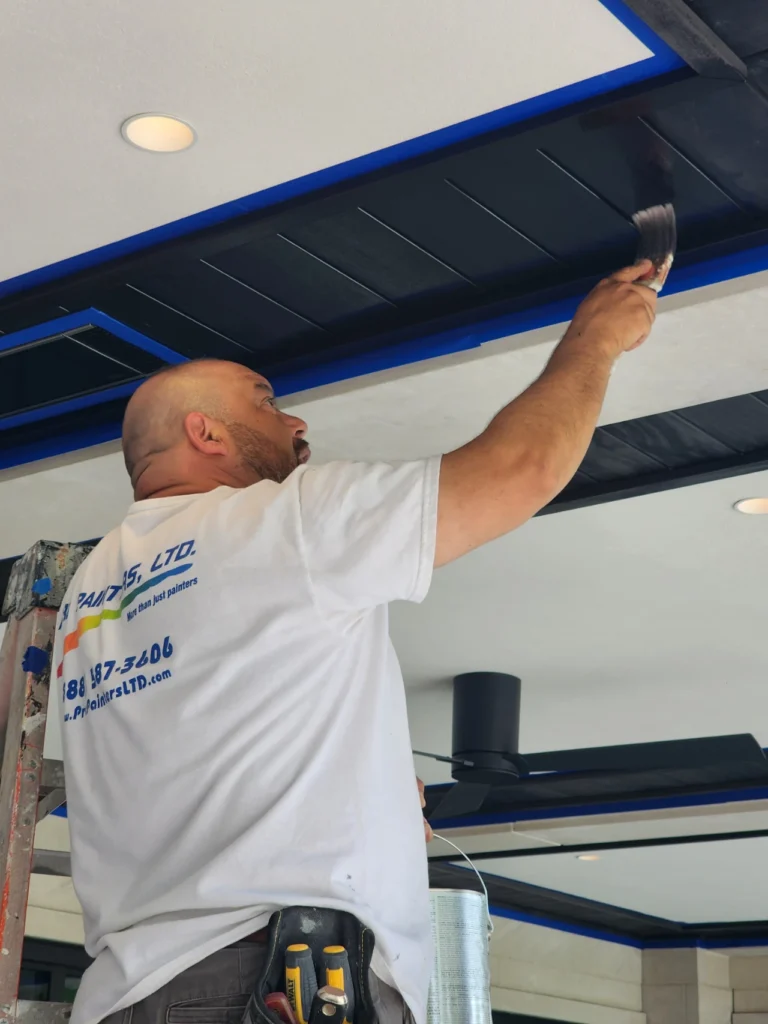 Pro Painters LTD crew member applying black paint to coffered ceiling soffit with blue trim, white ceiling in background