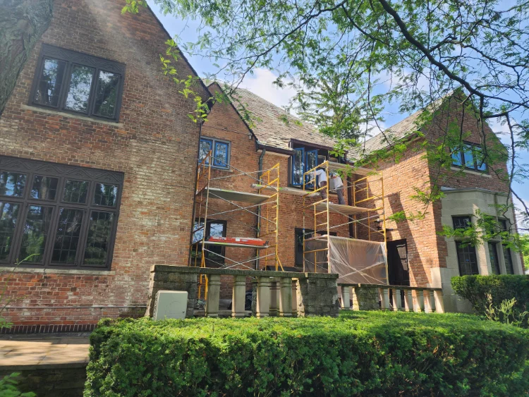 Exterior view of a two-story red brick house undergoing restoration, featuring yellow steel scaffolding with wooden platforms where Pro Painters LTD workers are working on open, blue-taped leaded glass dormer windows.