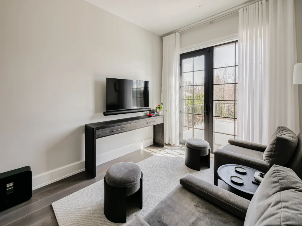 Modern living room with a wall-mounted TV, gray furniture, a console table, large window with white curtains, and a neutral color palette.