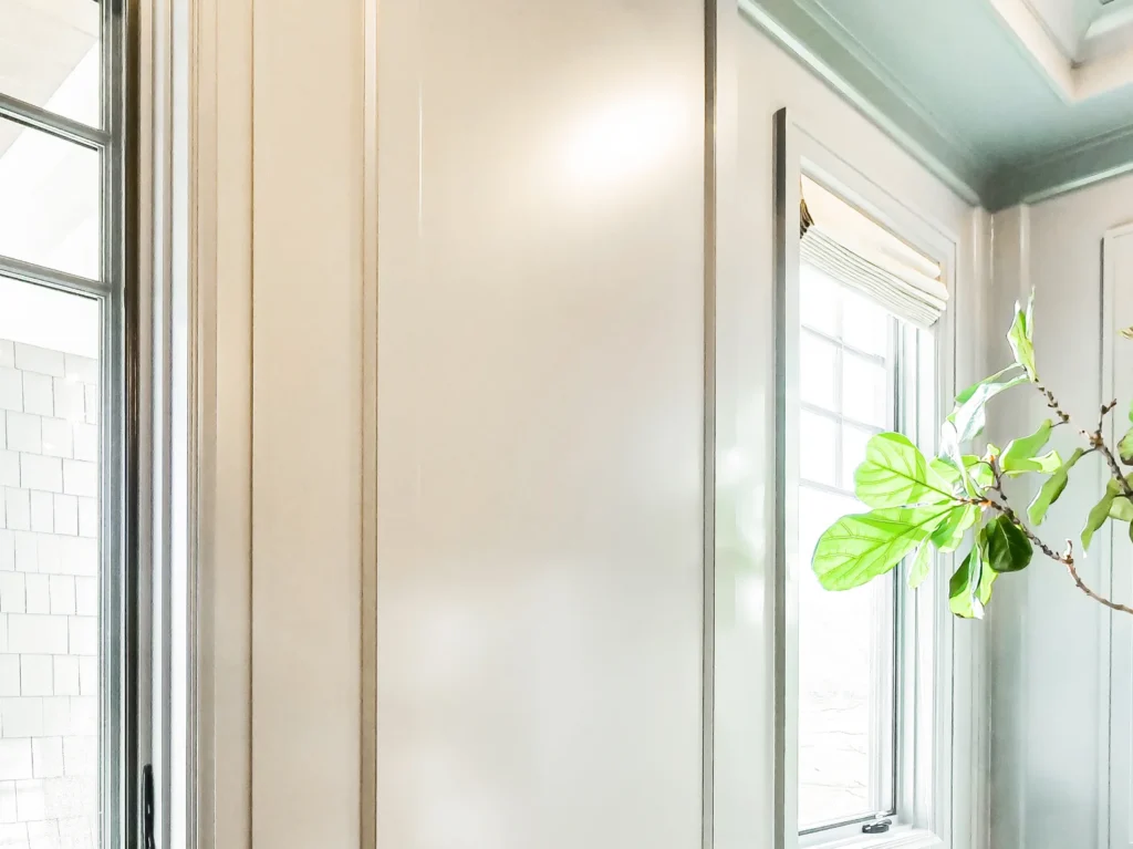 Close-up interior shot of pristine off-white millwork and wall paneling, featuring a bright double-hung window with a light Roman shade and green fiddle-leaf fig leaves softly catching natural sunlight