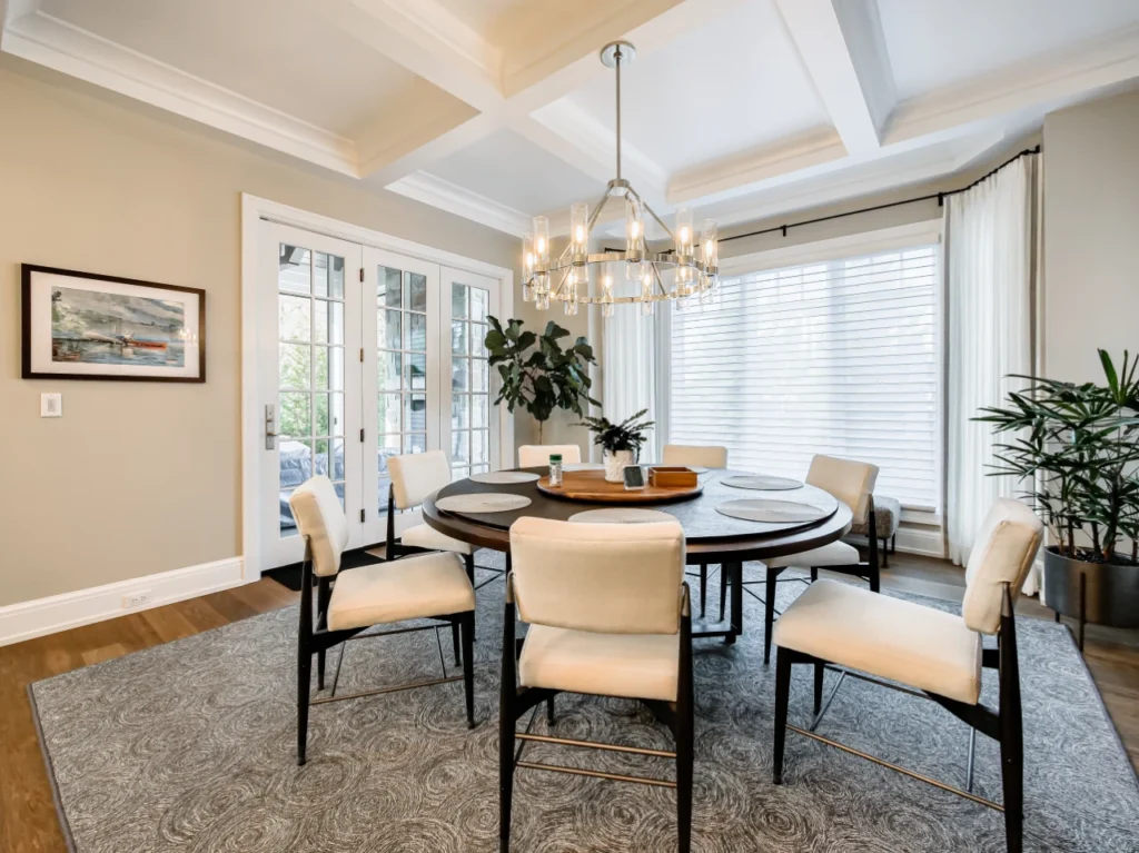 Formal dining space with a round dark wood dining table, surrounding cream upholstered chairs, a patterned grey area rug, coffered ceiling, modern glass cylinder chandelier, and french doors opening to an outdoor patio.