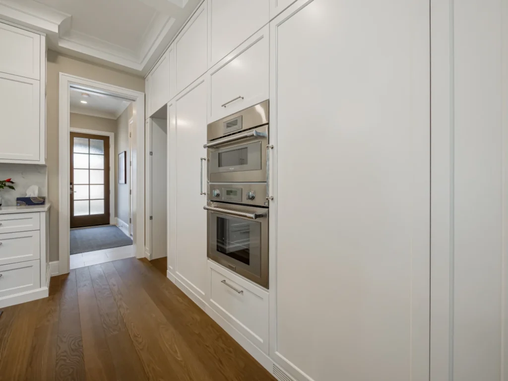Perspective view along a hallway showing integrated white floor-to-ceiling kitchen cabinetry housing built-in stainless steel wall ovens, leading toward a glass panel exterior door and rich hardwood flooring.