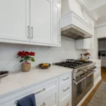 White custom kitchen cabinetry with quartz countertops, a stainless steel range and hood, and warm wood flooring.