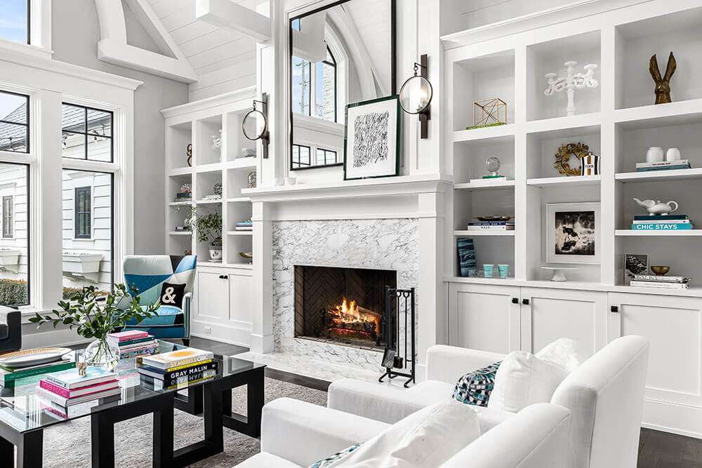Transitional great room in a Metro Detroit home with custom white built-in cabinetry, detailed painted millwork, vaulted ceiling beams, black-framed windows, marble fireplace surround, integrated display shelving, and crisp architectural trim finished by Pro Painters LTD.